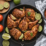 Grilled chicken legs arranged in a skillet, garnished with lime wedges and fresh cilantro, surrounded by tomatoes and other ingredients.